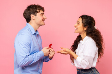 Young man proposing to his beloved lady, giving her engagement ring on Valentine's Day over pink studio background
