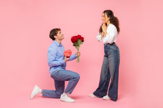 Romantic European guy giving his beloved girlfriend flowers and gift for Valentine's Day over pink studio background, full length shot