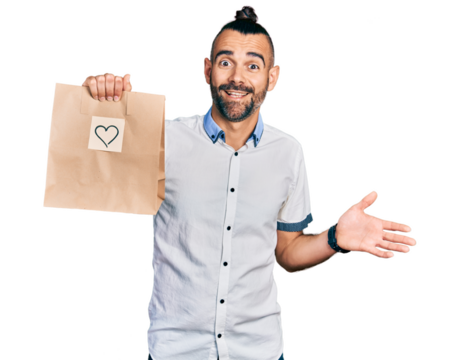 Hispanic man with ponytail holding take away paper bag with heart reminder celebrating achievement with happy smile and winner expression with raised hand