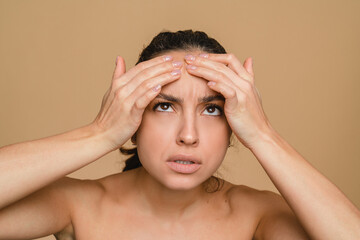 Acne pimples on face skin. Young hispanic sad woman looking for wrinkles on her face isolated in beige background. Anti-age cosmetics care treatment. Health dermatology skin problem.