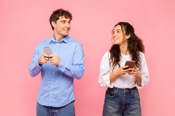 Happy European spouses using cellphones, looking at each other and smiling, texting online or surfing Internet, posing on pink studio background