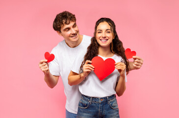 Happy loving European couple holding red heart-shaped cards, posing over pink studio background. St. Valentines day concept