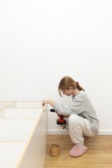 Naklejka premium A woman assembles furniture at home using an electric drill, showing independence and ability to perform repair work. The concept of DIY, home comfort and independent problem solving skills.