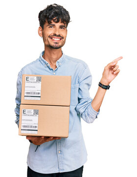Young hispanic man holding delivery package smiling happy pointing with hand and finger to the side