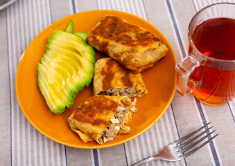 Pancakes stuffed with stewed liver and egg filling, toasted until golden brown. Pancakes are on plate with slices of ripe avocado fruit. Hearty breakfast, protein diet