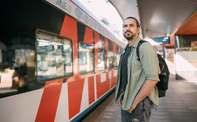 A man is waiting for a train on the platform. A young guy, a passenger with a backpack is standing on the platform waiting for the train