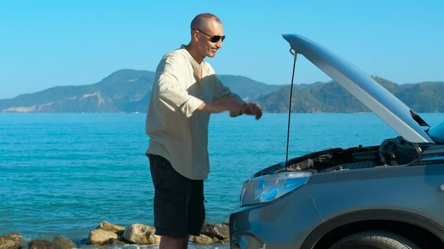 Problems with the car while traveling. Male tourist is having problems with his car on a beautiful sunny day by the sea. He looks stressed as he tries to fix the issue