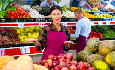 Polite friendly Asian saleswoman meeting customers in greengrocery store, offering fresh fruits and vegetables for sale