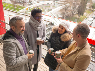A group of four business colleagues taking a coffee break outdoors on a winter day. They are standing together, smiling, and holding coffee cups in an urban business district. Dressed in warm coats