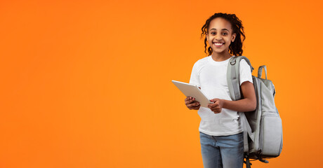 E-learning Concept. Portrait of smiling African American teenage girl holding and using digital tablet over yellow orange studio background, looking and posing at camera, wearing backpack