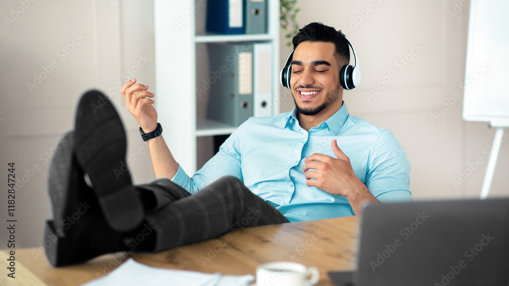 Canvas Prints Joyful Arab company employee listening to music in headphones and playing virtual guitar at desk in modern office. Millennial businessman having fun on break, relaxing at workplace