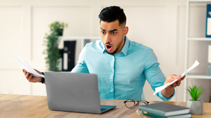 Shocked young Arab businessman with documents looking at laptop screen in dismay, shouting WTF, making mistake in project at office. Business crisis, error, deadline concept