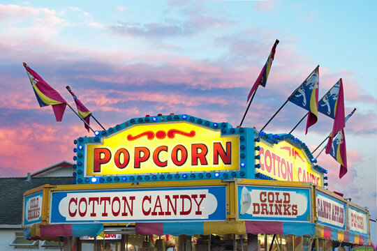 A fair concession that sells popcorn and cotton candy illuminated during a colorful sunset