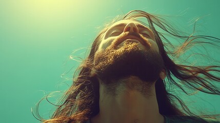 Man with long hair and beard looking up with eyes closed against a teal sky. Sunlight illuminates his face. Ideal for lifestyle or wellness campaigns.