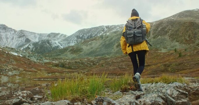 Female hiker with backpack carefully crossing stream on rocks near mountain lake with snowy peaks in the background, enjoying a challenging hike in autumn nature