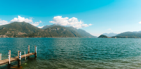 Tranquil scene of a wooden pier extending into the calm waters of Lake Como, surrounded by lush green mountains and a clear blue sky, creating a peaceful and scenic landscape in Menagg Lombardy, Italy
