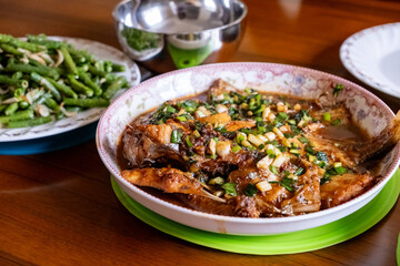 A Chinese home-cooked meal featuring a fish smothered in rich soy sauce, garnished with chopped scallions, and served with a side dish of stir-fried green beans.