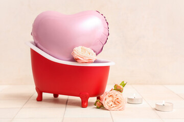 Mini bathtub with heart-shaped balloon, candles and roses on white tile near wall. Valentine's Day celebration