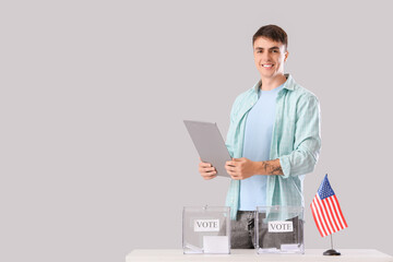 Voting young man with clipboard near ballot box and USA flag at table on light background