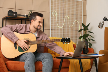 Young happy male composer with laptop playing guitar on sofa in music studio