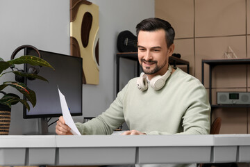Young happy male composer with headphones and notes playing synthesizer in music studio