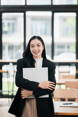 Young woman in a modern office holding a laptop, smiling confidently with large windows in the background.