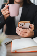 A business professional in a suit using a smartphone and drinking coffee at a desk with a notebook and laptop.