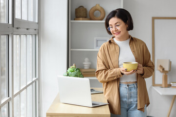 Mature woman with cup of tea and laptop on commode at home