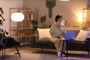 Mature woman with cup of tea and laptop video chatting on sofa at home in evening