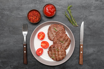 Plate with tasty meat cutlets, cut tomatoes, rosemary, bowl of ketchup and cutlery on black grunge background