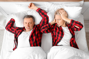 Displeased mature woman lying with her snoring husband on bed, top view