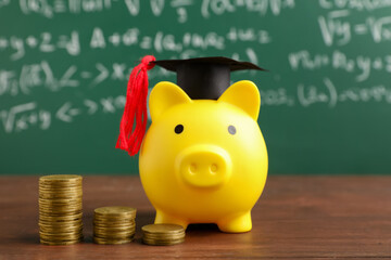 Piggy bank with graduation hat and coins on table near green blackboard. Student loan concept