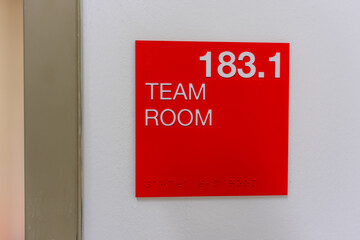 Red and white team room sign, 183.1, with braille code on a white interior building wall.	
