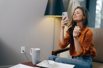 Young woman enjoying a healthy snack while using her smartphone, seated comfortably in a cozy modern living room with soft lighting and warm colors