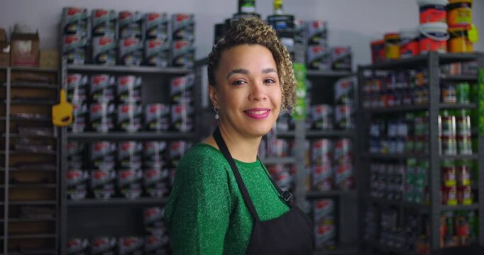 Confident woman smiling in local small business hardware store, standing in front of shelves stocked with paint and tools, showcasing professionalism and a welcoming demeanor