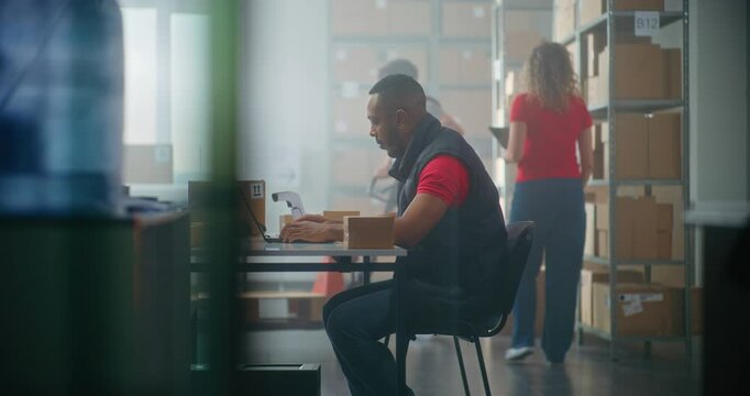 African American post office worker works on laptop computer, monitors parcels information. Sorting center workers checking cardboard boxes for delivery to clients. Online shop warehouse. Slow motion.
