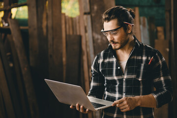 Young Caucasian male worker looking at laptop computer in wood workshop for review design project.