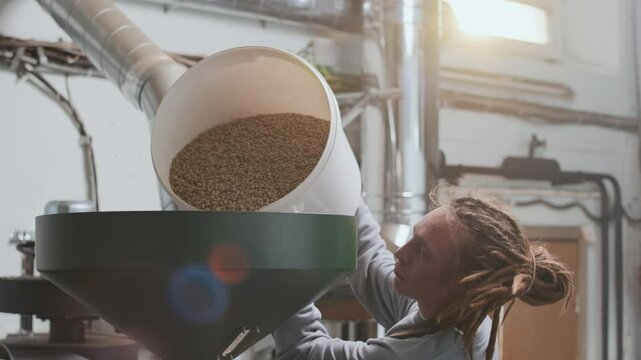 Tilt up shot of male Caucasian technician with dreadlocks adding coffee beans to professional grinding machine at factory