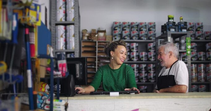 Man and woman conversing casually at counter of local small business hardware store, surrounded by shelves of paint and tools, friendly workplace interaction between colleagues
