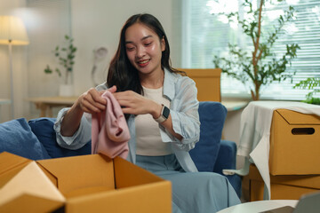 Smiling young asian woman unpacking clothes from a cardboard box while sitting on the sofa in her...