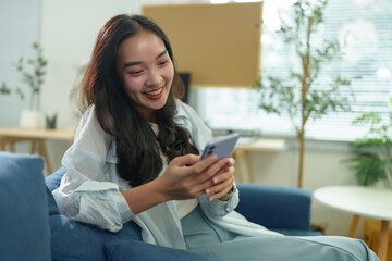 Happy millennial woman using a smartphone while relaxing on a comfortable sofa in her living room, enjoying online communication and social media