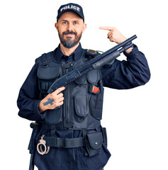 Young handsome man wearing police uniform holding shotgun pointing finger to one self smiling happy...
