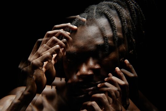 Distorted image with double exposure effect of Black man posing with hands on face looking at camera with deep shadows covering his face in dark in studio, copy space