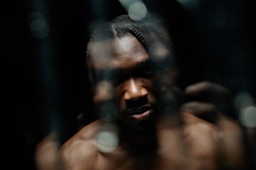 Shot through glass with water running down of Black man staring at camera with scary look hidden in deep shadows casting from top contrast light in studio with black background, copy space © Seventyfour