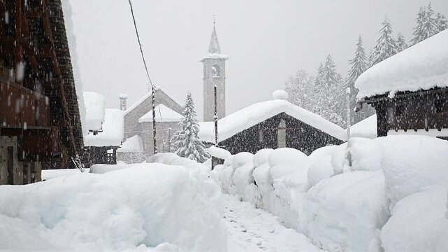 Wooden huts of a traditional mountain village in the European Alps during a heavy snowfall with a church and bell tower in the background. Macugnaga village, important ski resort in northern Italy