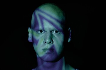 Portrait of emotionless black eyed man with bald head looking at camera with piercing stare while standing in blue light of projector in darkness, copy space