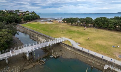 Fototapeta premium Coastal park scene with people enjoying the day in Milford. Picnic blankets, walkers, and a bridge over the waterway. Beautiful day at the park. Milford, Auckland, New Zealand