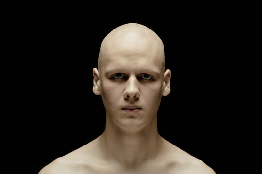 Portrait of unemotional pale man with with bald head staring at camera with scary gaze standing in hard light against black background in studio amplifying chilling atmosphere, copy space