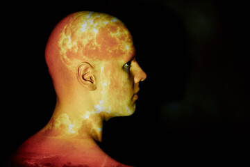 Profile shot of emotionless bald man with image of nuclear explosion flame projected onto face posing against black background in studio, copy space