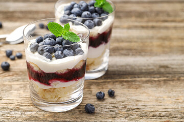Tasty trifle dessert. Sponge cake, blueberries, jam and whipped cream in glasses on wooden table, closeup. Space for text